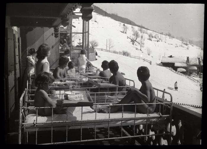 Enfants allités en cure de soleil et d'air pur sur une véranda. Des infirmières prennent soin d'eux.